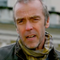 A imagem apresenta um close do ator britânico John Hannah. Ele aparece com uma expressão levemente confusa ou franzida, olhando diretamente para a câmera.