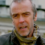 A imagem apresenta um close do ator britânico John Hannah. Ele aparece com uma expressão levemente confusa ou franzida, olhando diretamente para a câmera.