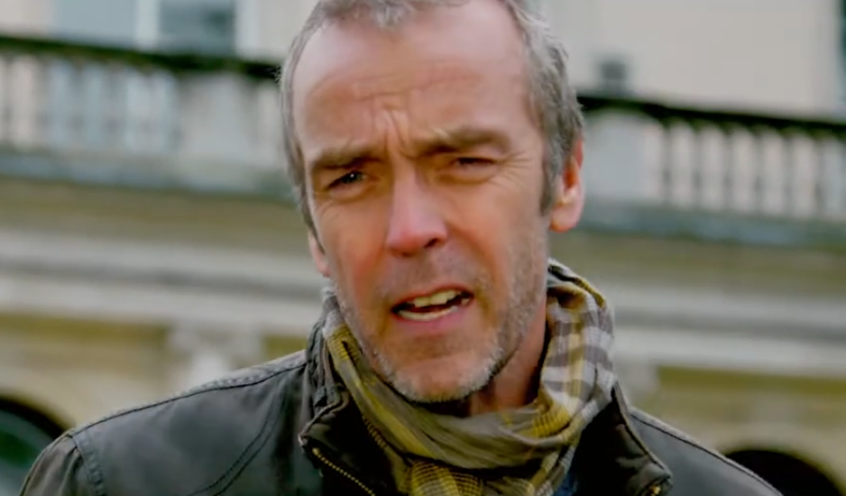 A imagem apresenta um close do ator britânico John Hannah. Ele aparece com uma expressão levemente confusa ou franzida, olhando diretamente para a câmera.