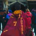 Harry Potter caminhando em direção ao campo de Quadribol de Hogwarts usando capa da Grifinória