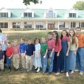 A família Duggar reunida em frente a uma grande casa branca com telhado verde em uma área gramada.