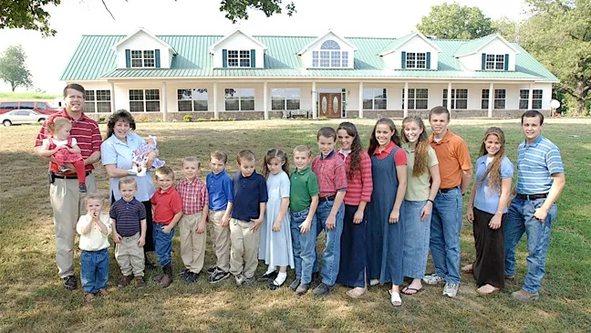 A família Duggar reunida em frente a uma grande casa branca com telhado verde em uma área gramada.
