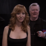 Cena da série The Comeback em formato de falso documentário. Em primeiro plano, Lisa Kudrow como Valerie Cherish, com cabelos ruivos ondulados e um vestido preto de alças. Logo atrás dela, à direita, o ator Robert Michael Morris como Mickey Deane, vestindo uma camisa preta, óculos pendurados no pescoço e vários anéis grandes nos dedos. O fundo é escuro, sugerindo um ambiente de bastidores de produção.