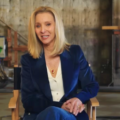 A atriz Lisa Kudrow sentada em uma cadeira de diretor em um set de filmagem, vestindo um blazer de veludo azul escuro sobre uma camisa branca e usando um colar com pingente vermelho. Ela sorri para a câmera em um ambiente de bastidores com equipamentos de estúdio ao fundo.