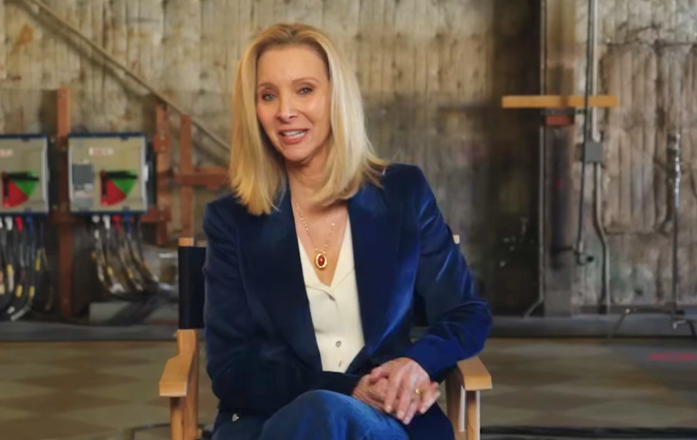 A atriz Lisa Kudrow sentada em uma cadeira de diretor em um set de filmagem, vestindo um blazer de veludo azul escuro sobre uma camisa branca e usando um colar com pingente vermelho. Ela sorri para a câmera em um ambiente de bastidores com equipamentos de estúdio ao fundo.