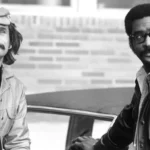 Uma fotografia em preto e branco mostrando dois homens ao lado de um carro. Ed Bernard, à direita, usa óculos e jaqueta de couro preta. O homem à esquerda usa boina e jaqueta clara.