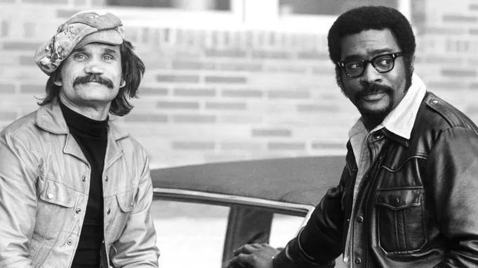 Uma fotografia em preto e branco mostrando dois homens ao lado de um carro. Ed Bernard, à direita, usa óculos e jaqueta de couro preta. O homem à esquerda usa boina e jaqueta clara.