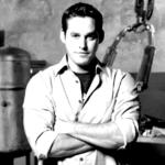 Retrato do ator Nicholas Brendon sorrindo, com cabelos castanhos curtos e usando uma camisa social clara. O fundo é neutro, destacando a expressão do artista.