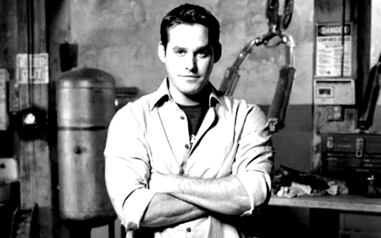 Retrato do ator Nicholas Brendon sorrindo, com cabelos castanhos curtos e usando uma camisa social clara. O fundo é neutro, destacando a expressão do artista.