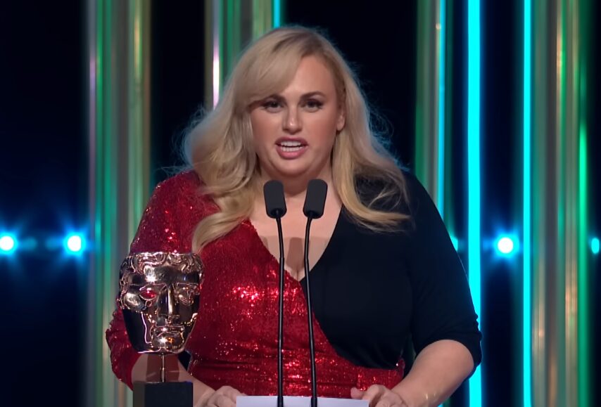 A atriz Rebel Wilson em um tapete vermelho sorrindo para fotógrafos.