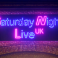 Membros do elenco do Saturday Night Live UK caminham por rua de Londres sob luzes de neon com o nome do programa.