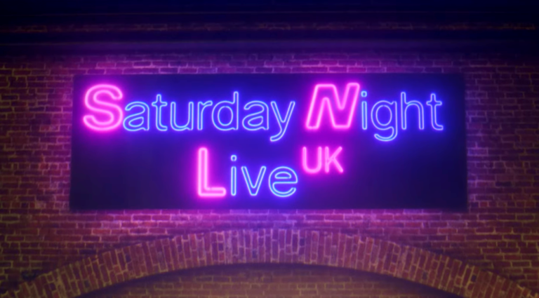 Membros do elenco do Saturday Night Live UK caminham por rua de Londres sob luzes de neon com o nome do programa.