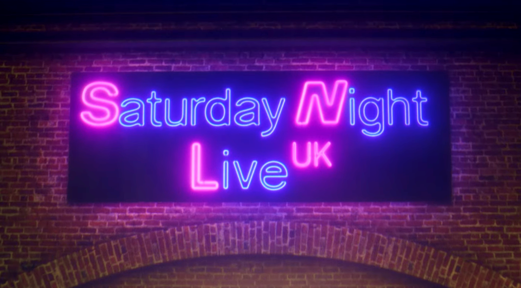 Membros do elenco do Saturday Night Live UK caminham por rua de Londres sob luzes de neon com o nome do programa.