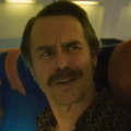 Um plano médio do ator Sam Rockwell com bigode e camisa estampada dentro de um avião comercial. Ele está olhando para a esquerda com uma expressão confusa. Outros passageiros e poltronas azuis e laranjas são visíveis ao fundo.