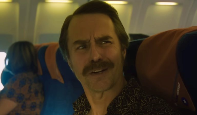 Um plano médio do ator Sam Rockwell com bigode e camisa estampada dentro de um avião comercial. Ele está olhando para a esquerda com uma expressão confusa. Outros passageiros e poltronas azuis e laranjas são visíveis ao fundo.