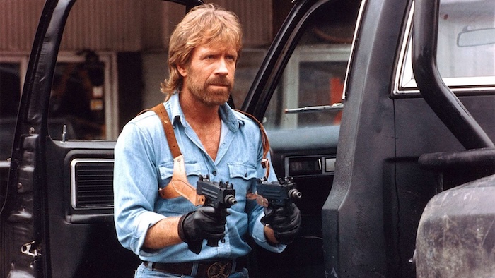 Chuck Norris segurando duas submetralhadoras Uzi e usando um colete de coldres sobre a camisa jeans.