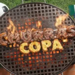 Tiago Medeiros sorri ao lado de uma churrasqueira em preparação para a Copa