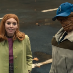 Natasha Lyonne, com cabelos ruivos e casaco verde, ao lado do Coisa (com boné azul e casaco), ambos olhando para cima em uma rua escura.