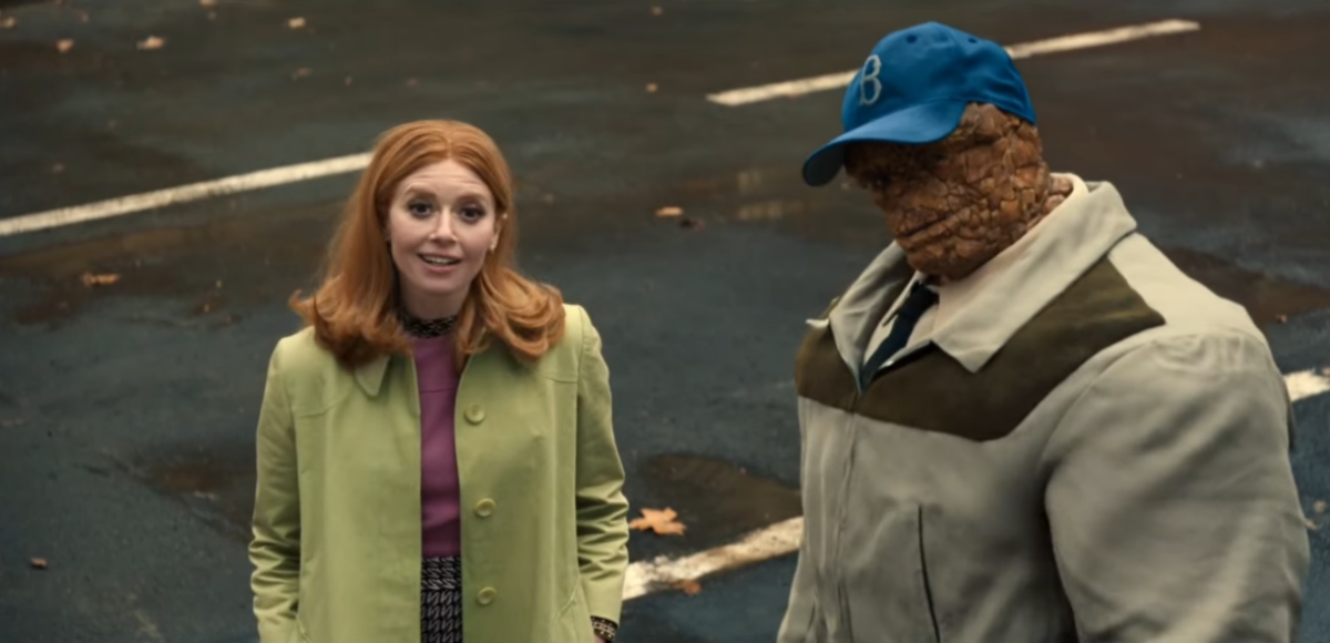 Natasha Lyonne, com cabelos ruivos e casaco verde, ao lado do Coisa (com boné azul e casaco), ambos olhando para cima em uma rua escura.