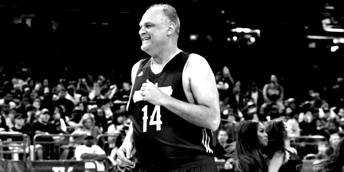 Oscar Schmidt sorrindo durante um evento esportivo, vestindo uma camisa polo escura.