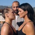Foto de perfil de Ronda Rousey e Gina Carano em um "face-off" (encarada de luta), olhando firmemente uma para a outra. Um homem careca está de pé entre elas, ao fundo. O cenário é um terraço com vista para colinas e uma cidade ao sol.