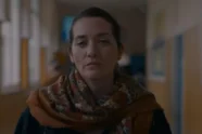 Atriz Selma Alaoui em cena do filme Aquela Noite, usando um cachecol estampado e encarando a câmera com expressão séria em um corredor subexposto.