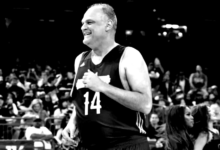 Oscar Schmidt sorrindo durante um evento esportivo, vestindo uma camisa polo escura.