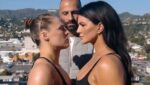 Foto de perfil de Ronda Rousey e Gina Carano em um "face-off" (encarada de luta), olhando firmemente uma para a outra. Um homem careca está de pé entre elas, ao fundo. O cenário é um terraço com vista para colinas e uma cidade ao sol.
