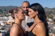 Foto de perfil de Ronda Rousey e Gina Carano em um "face-off" (encarada de luta), olhando firmemente uma para a outra. Um homem careca está de pé entre elas, ao fundo. O cenário é um terraço com vista para colinas e uma cidade ao sol.