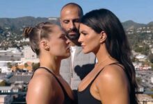 Foto de perfil de Ronda Rousey e Gina Carano em um "face-off" (encarada de luta), olhando firmemente uma para a outra. Um homem careca está de pé entre elas, ao fundo. O cenário é um terraço com vista para colinas e uma cidade ao sol.