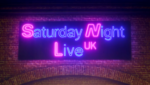 Membros do elenco do Saturday Night Live UK caminham por rua de Londres sob luzes de neon com o nome do programa.