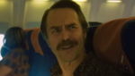 Um plano médio do ator Sam Rockwell com bigode e camisa estampada dentro de um avião comercial. Ele está olhando para a esquerda com uma expressão confusa. Outros passageiros e poltronas azuis e laranjas são visíveis ao fundo.